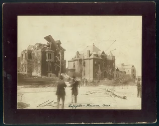 Foto horizontal en sepia mostrando tres casas parcialmente destruidas, árboles despojados y espectadores mirando los daños causados por un tornado